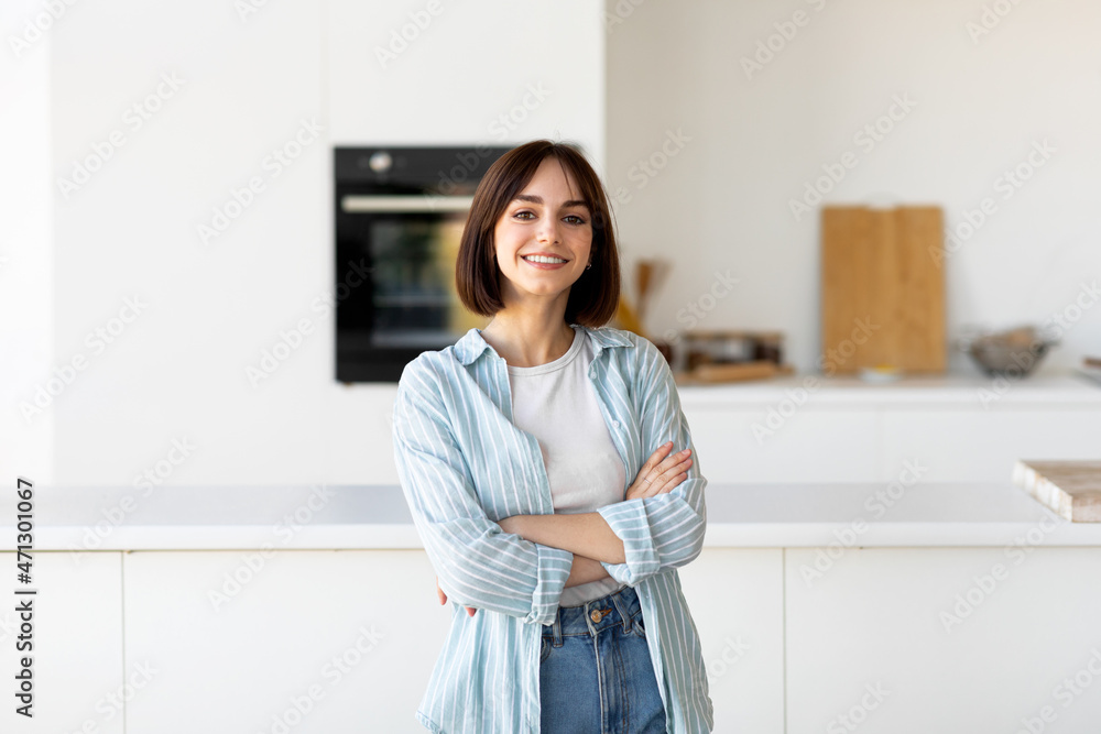Happy person. Portrait of young lady posing with crossed hands, smiling at camera, standing in modern kitchen interior Happy person. Portrait of young lady posing with crossed hands, smiling at camera, standing in modern kitchen interior
