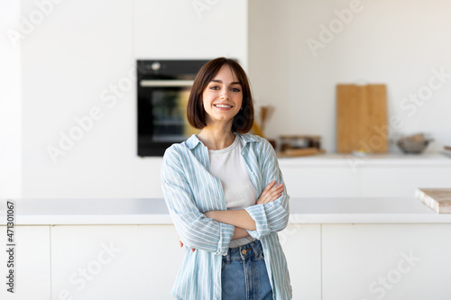 Happy person. Portrait of young lady posing with crossed hands, smiling at camera, standing in modern kitchen interior
