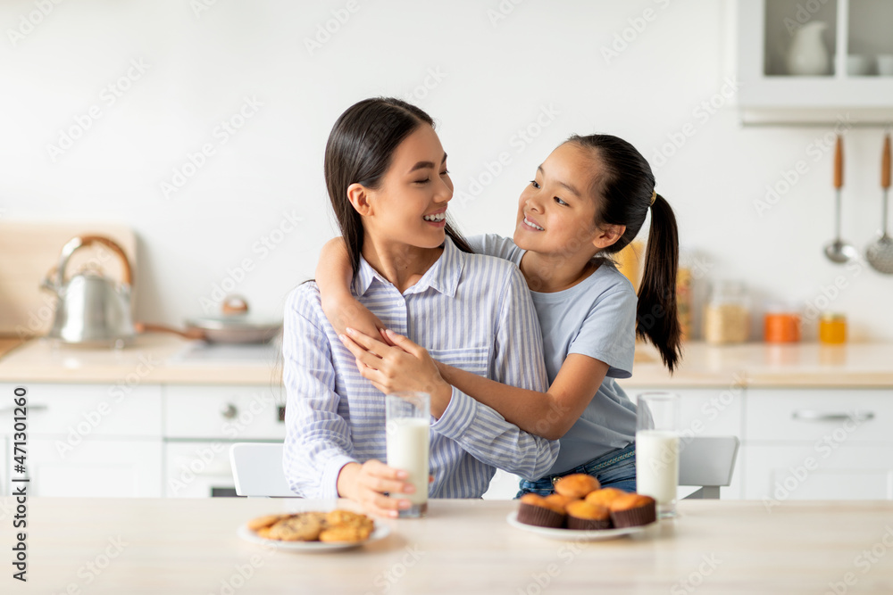 Cute asian daughter hugging her young happy mom while enjoying homemade cookies and biscuits in kitchen