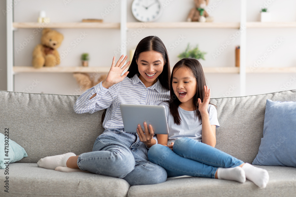 Modern remote communication concept. Excited asian mother and daughter having video call on tablet, sitting on sofa Modern remote communication concept. Excited asian mother and daughter having video call on tablet, sitting on sofa