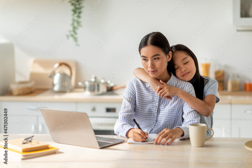 Family business. Young asian woman working on laptop and taking notes, cute daughter embracing mother at home Family business. Young asian woman working on laptop and taking notes, cute daughter embracing mother at home