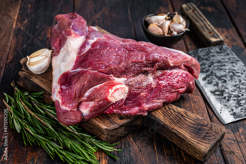 Raw goat thigh on butcher board with meat cleaver. Dark wooden background. Top view. Copy space