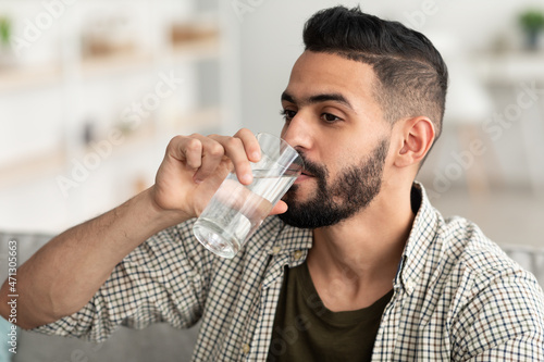 Healthy lifestyle concept. Millennial Arab guy drinking water from glass at home