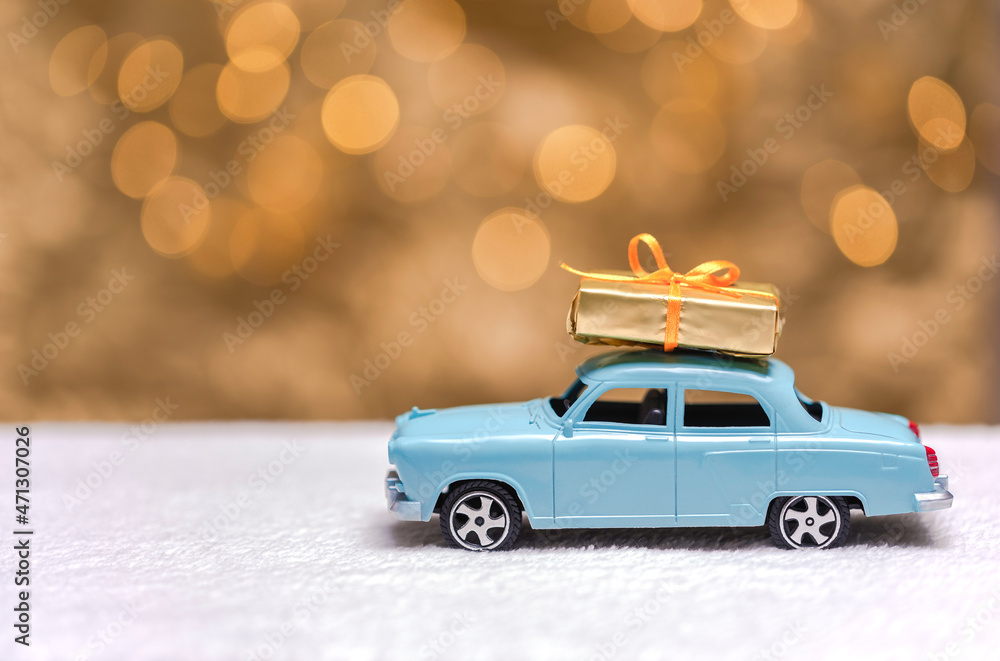 Small blue toy car with gift box on the roof. Golden festive background with bokeh lights at the background