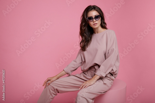 High fashion photo of beautiful elegant young woman in a pretty pink jumpsuit, sunglasses posing on studio background. Slim figure. Model sits on cube cylinder. Monochrome. Luxurious hair, shiny curls