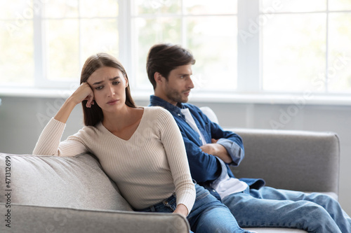 Sad depressed young caucasian wife ignores unhappy husband sitting on sofa in living room interior