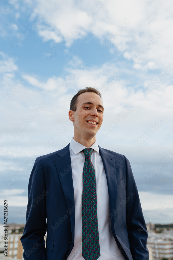 Young businessman in a suit and tie looking at camera and smiling while standing outside in a rooftop with a cloudy sky Young businessman in a suit and tie looking at camera and smiling while standing outside in a rooftop with a cloudy sky
