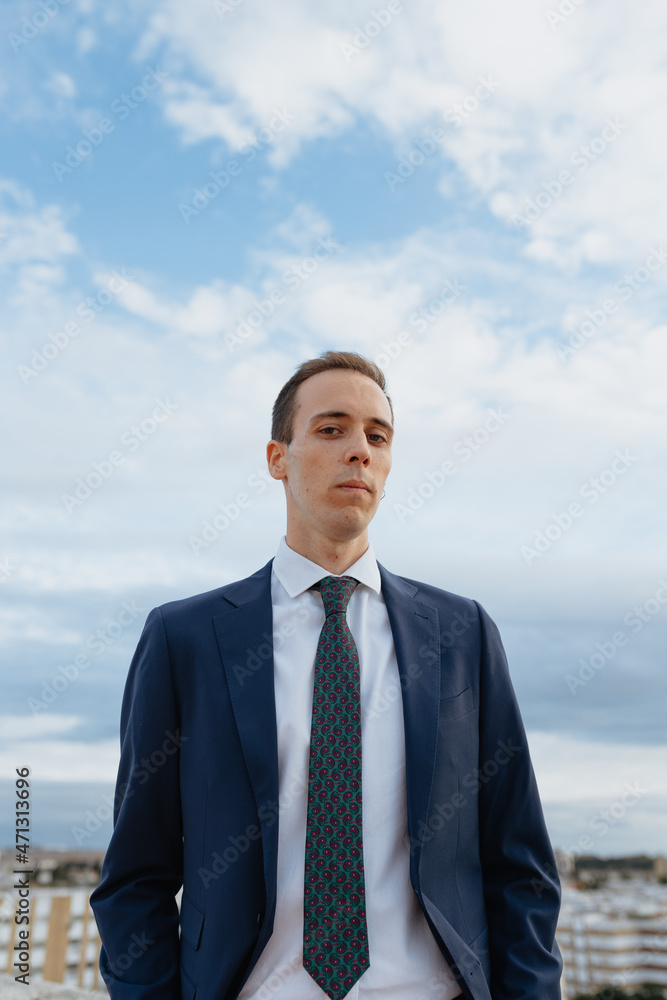 Young businessman in a suit and tie looking serious at camera standing outside in a rooftop with a cloudy sky Young businessman in a suit and tie looking serious at camera standing outside in a rooftop with a cloudy sky