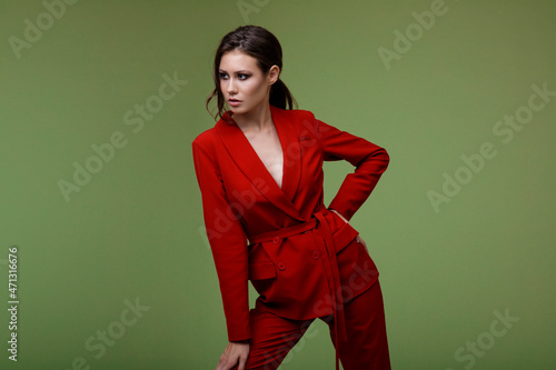 High fashion photo of a beautiful elegant young woman in a pretty red suit, jacket, pants, trousers posing over green background. Slim figure. Studio Shot. Businesswoman. Femininity and success