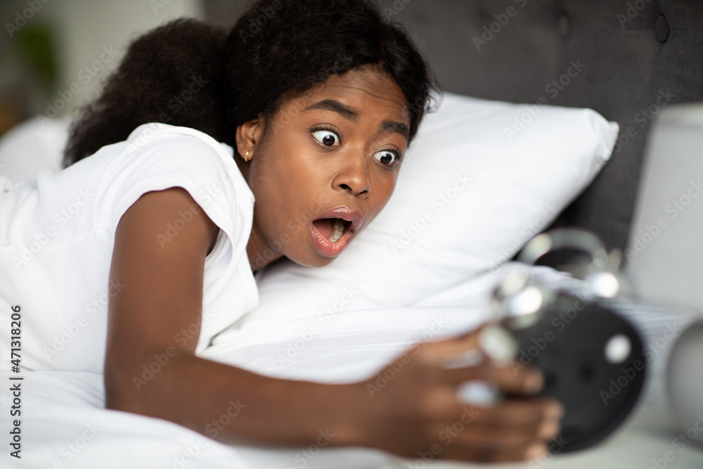 Woman hold big black clock on bed in bedroom at home. She shock after wake up too late for appointment or work or class study. - obrazy, fototapety, plakaty 
