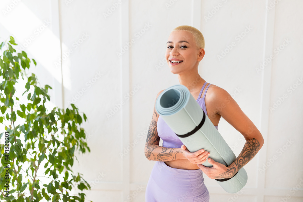 Portrait of positive woman holding yoga mat and smiling at camera while standing against white wall, ready for training Portrait of positive woman holding yoga mat and smiling at camera while standing against white wall, ready for training
