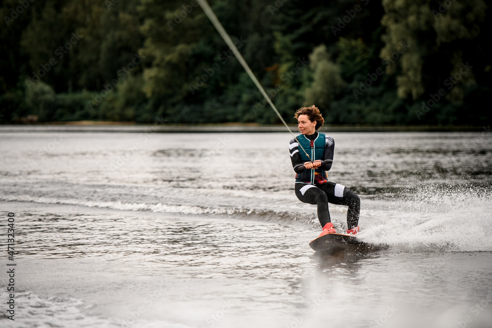 view of young woman in wetsuit riding on river water on wakeboard holding rope with her hands view of young woman in wetsuit riding on river water on wakeboard holding rope with her hands
