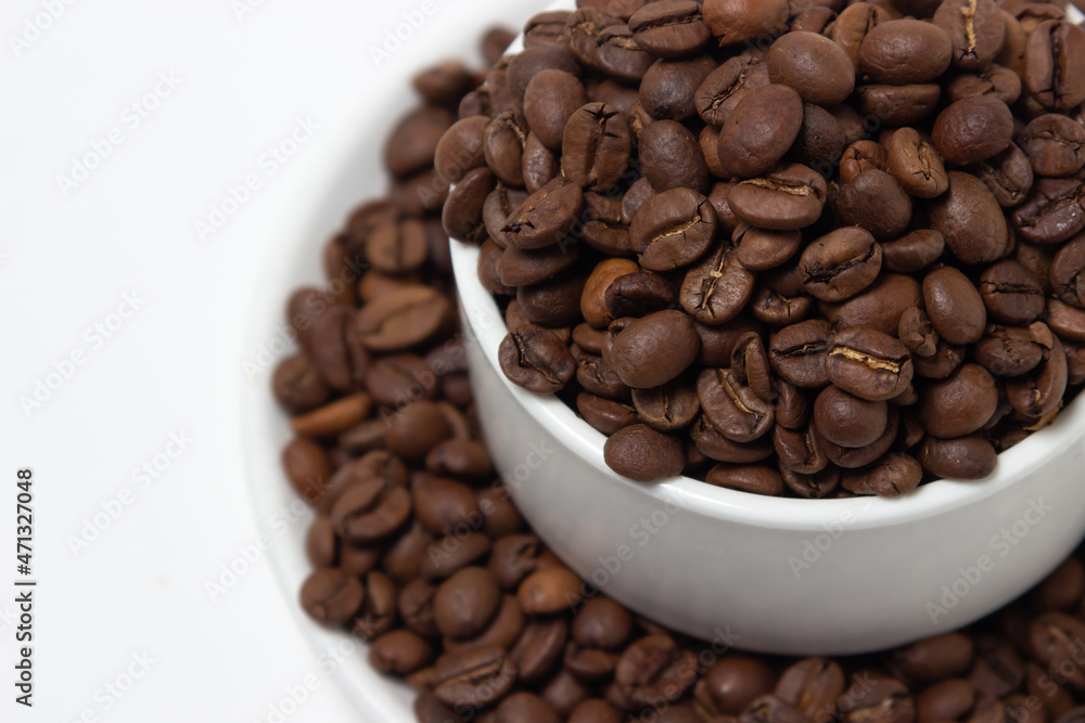 Fototapeta premium White cup overflowing with coffee beans on a white background. Freshly roasted coffee beans