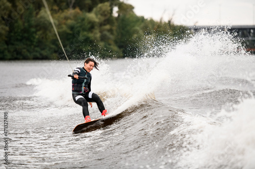 beautiful cheerful girl in wetsuit actively rides waves on wakeboard on the river