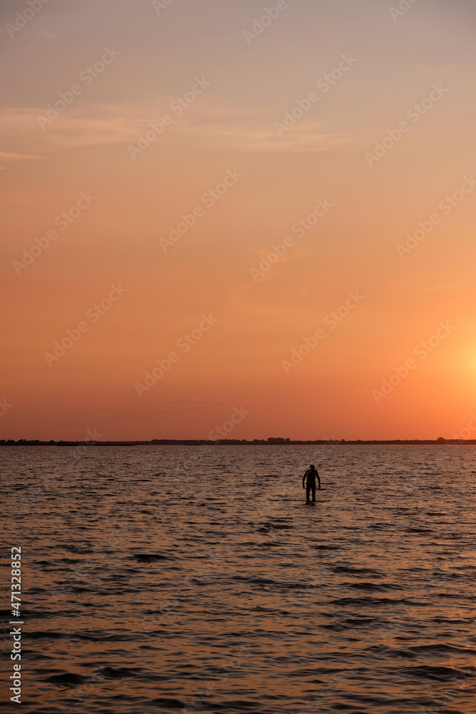 Silhouette of a lonely man in the water on a coral color sunset background. Svityaz lake sunset. Vertical image. 