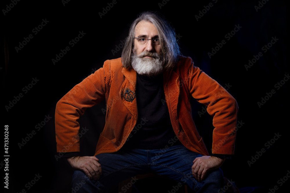 Portrait of a mature man in a stylish orange jacket on a black background . A fashionable intellectual looks into the camera with mockpice and interest. 