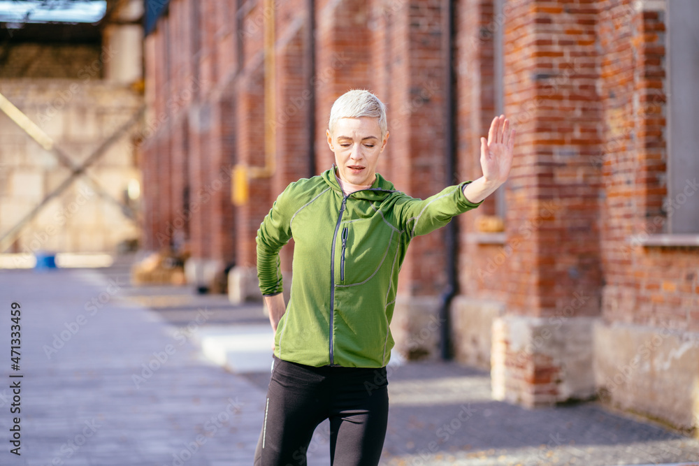 Preparation for running workout. Portrait of charming fit slim blond woman stretching legs enjoying the sun, female runner doing exercises against street wall background.