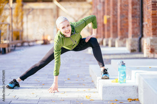 Contemporary middle age blonde female with short hair in activewear doing exercise for leg stretch while enjoying workout in urban environment. photo