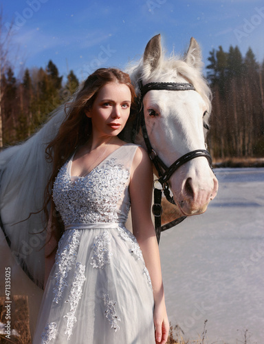 Beautiful young woman in grey sleeveless dress standing with white horse near winter lake. Fairytale portrait of a girl. Winter fantasy