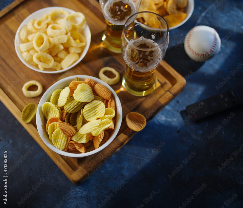 Classic Sports Fan Kit - Light beer in glasses, potato chips, onion rings on a wooden tray, ball and TV remote. Rest, relaxation, watching sports games on TV with friends.