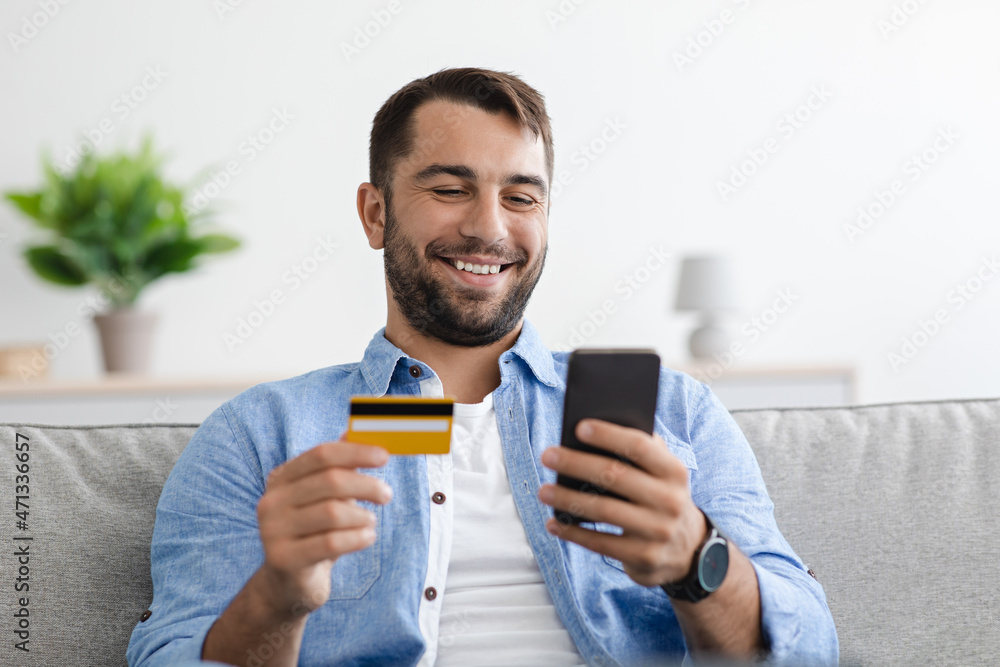 Smiling satisfied adult caucasian man with beard holds credit card and pays money online banking on smartphone Smiling satisfied adult caucasian man with beard holds credit card and pays money online banking on smartphone