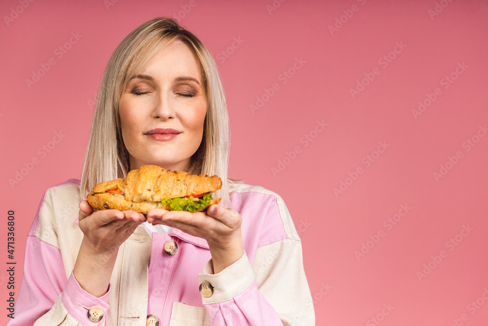 Portrait of young beautiful hungry blonde woman eating croissant sandwich. Isolated portrait of woman with fast food over pink background. Diet concept. Portrait of young beautiful hungry blonde woman eating croissant sandwich. Isolated portrait of woman with fast food over pink background. Diet concept.