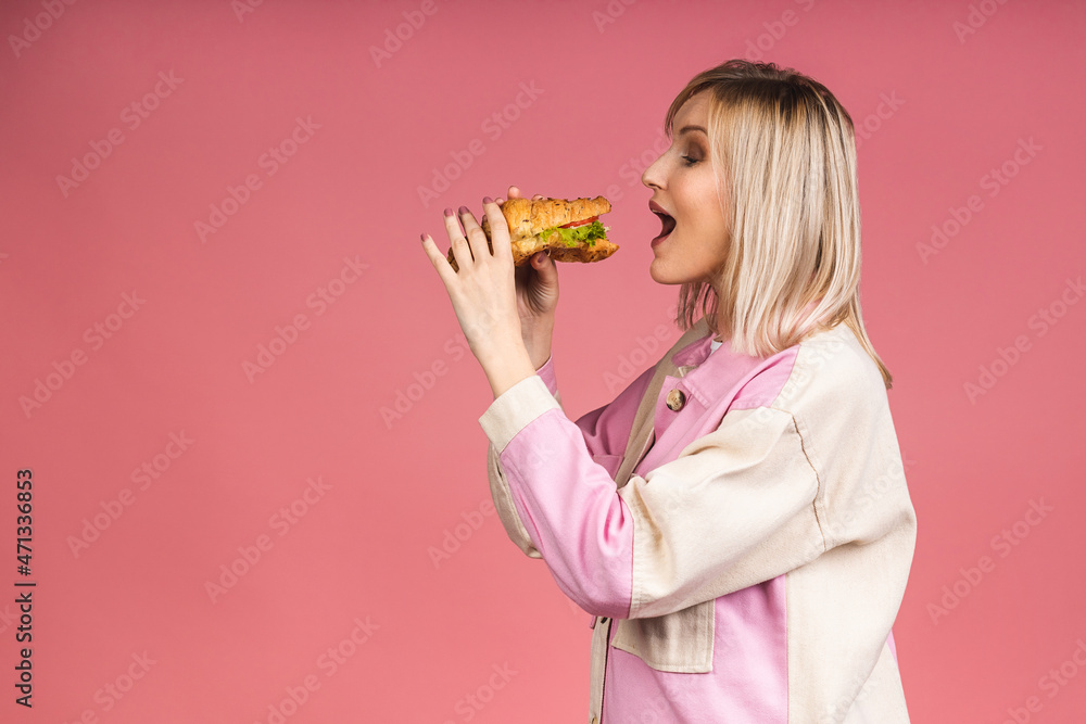 Portrait of young beautiful hungry woman eating croissant sandwich. Isolated portrait of woman with fast food over pink background. Diet concept. Portrait of young beautiful hungry woman eating croissant sandwich. Isolated portrait of woman with fast food over pink background. Diet concept.