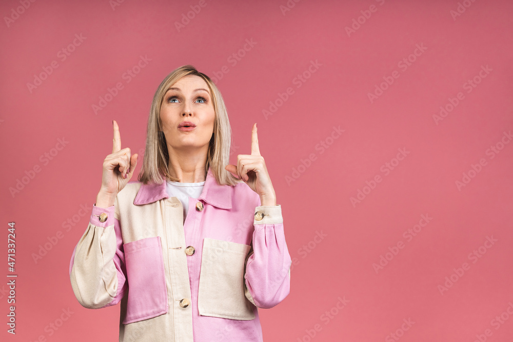 Portrait of a young beautiful cute cheerful blonde woman smiling looking at the camera isolated over pink background. Pointing fingers up. Portrait of a young beautiful cute cheerful blonde woman smiling looking at the camera isolated over pink background. Pointing fingers up.