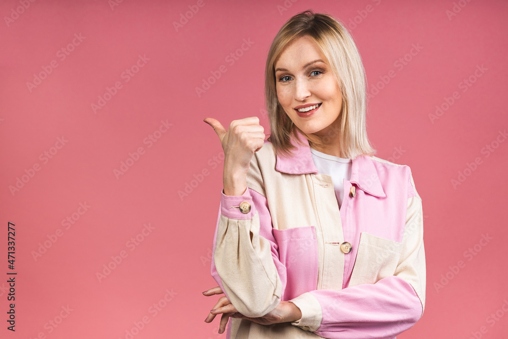 Portrait of a young beautiful cute cheerful blonde woman smiling looking at the camera isolated over pink background. Pointing finger away.