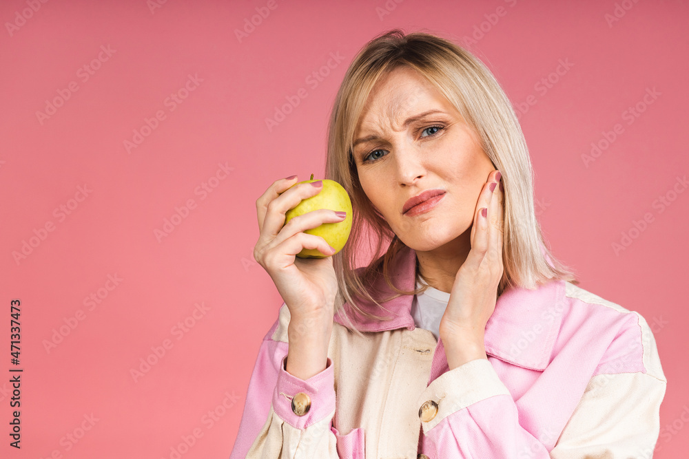 Young blonde woman touching mouth with hand with painful expression because of toothache or dental illness on teeth holding green apple. Dentist concept isolated over pink background. Young blonde woman touching mouth with hand with painful expression because of toothache or dental illness on teeth holding green apple. Dentist concept isolated over pink background.