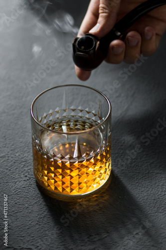 Smoking pipe in hand and a glass of whiskey on a dark table 
