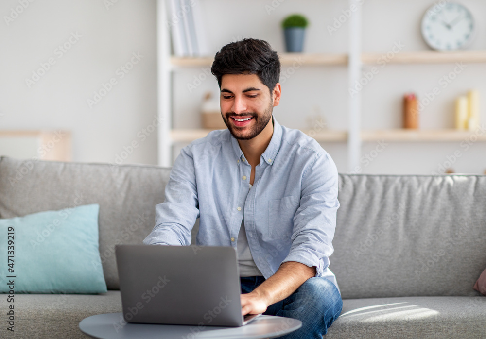 Home office concept. Happy arab guy working on pc laptop, sitting on comfy sofa at home interior, free space Home office concept. Happy arab guy working on pc laptop, sitting on comfy sofa at home interior, free space