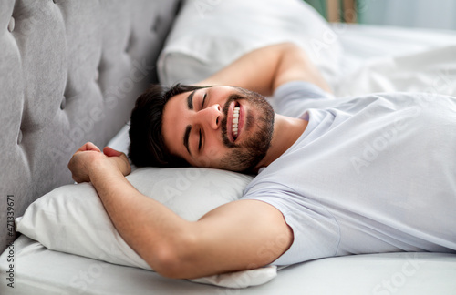 Happy wellslept arab man waking up in morning, stretching and smiling with closed eyes while lying in comfortable bed