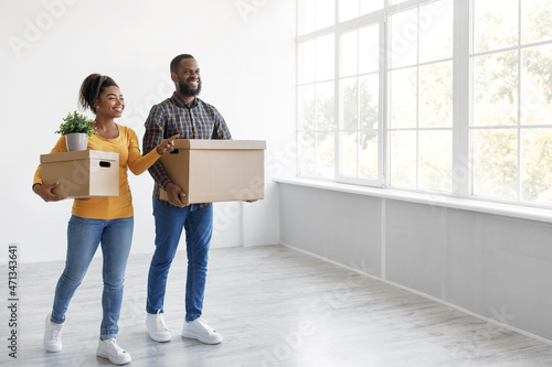 Smiling young african american husband and wife carry cardboard boxes and plant, look at empty room and window