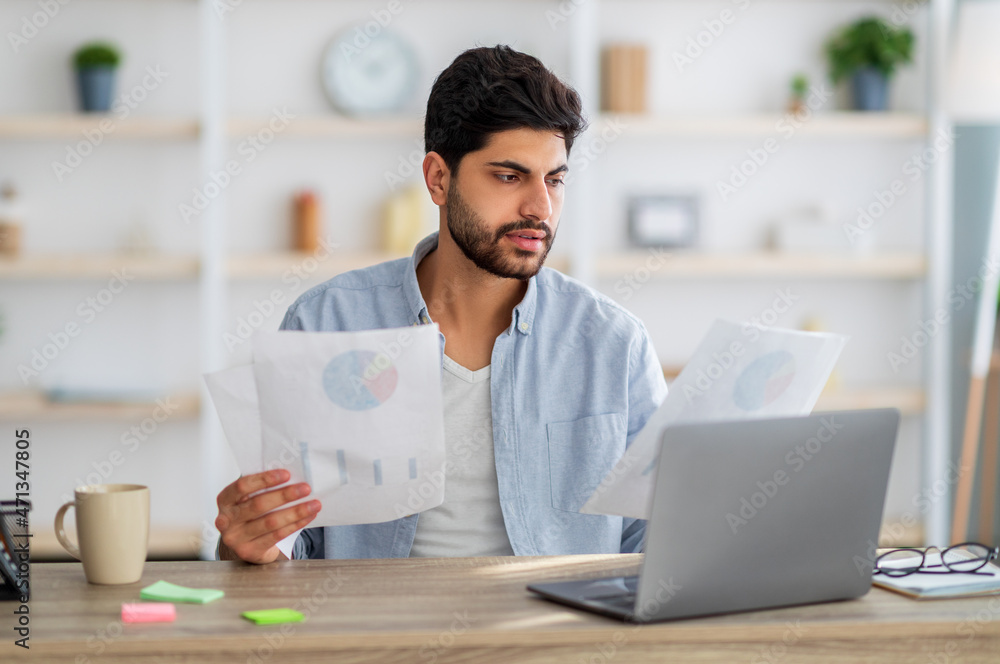 Serious arab guy looking through documents, having online business meeting on laptop while sitting at home office Serious arab guy looking through documents, having online business meeting on laptop while sitting at home office