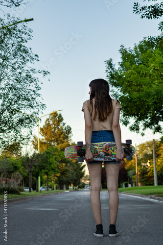 Woman standing on her back, holding her skateboard with both hands, looking to the side. On a city street