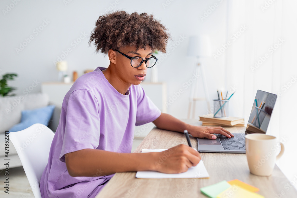 Focused black teenager taking notes during online lecture on laptop at home, free space Focused black teenager taking notes during online lecture on laptop at home, free space