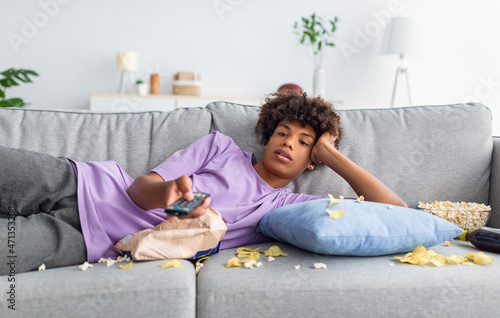 Fotografie Lazy black teen guy lying on sofa with scattered food, holding TV controller, wa