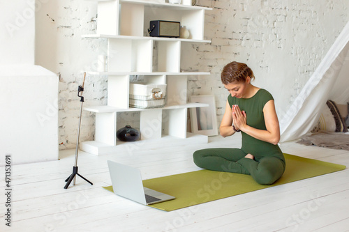 A beautiful woman in a green sports jumpsuit, in a bright room, is broadcasting online, performs a yoga exercise