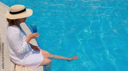 luxury senior woman dangling her legs in an outdoor swimming pool holding a blue refreshing cocktail wearing a straw hat. Summer rest. All inclusive