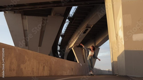 Extreme hobby and street fashion concept: trendy young female skating on longboard wearing denim shirt, white top and hipster sunglasses at sunset. Carefree girl with long curly hair longboarding photo