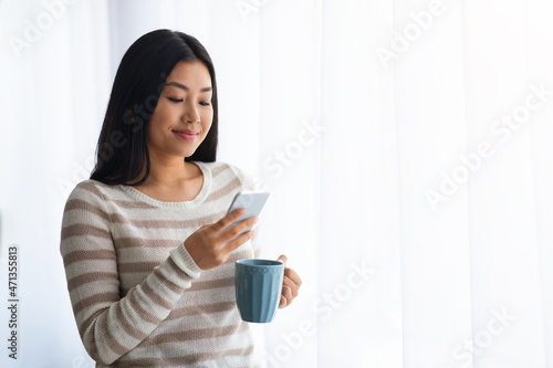 Young Asian Woman Using Smartphone And Drinking Coffee Near Window At Home