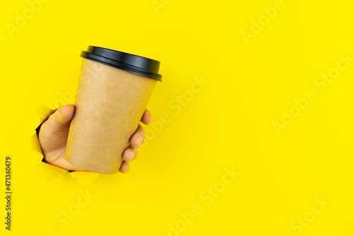 Simply minimal design. Female hand is holding paper coffee cup in torn hole of yellow colorful background. Takeaway drink and breakfast beverage. Good morning wake up awake concept. Mockup banner.