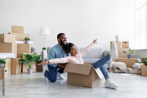 Smiling young african american husband pushes box with wife, rolls around living room with having fun, unpacking things