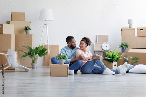 Cheerful millennial african american lady and guy hugging, sitting on floor with cardboard boxes © Prostock-studio