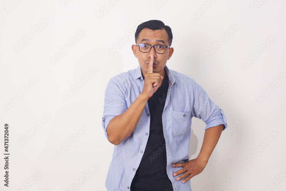Asian man over isolated white wall showing a sign of silence gesture putting finger in mouth Asian man over isolated white wall showing a sign of silence gesture putting finger in mouth