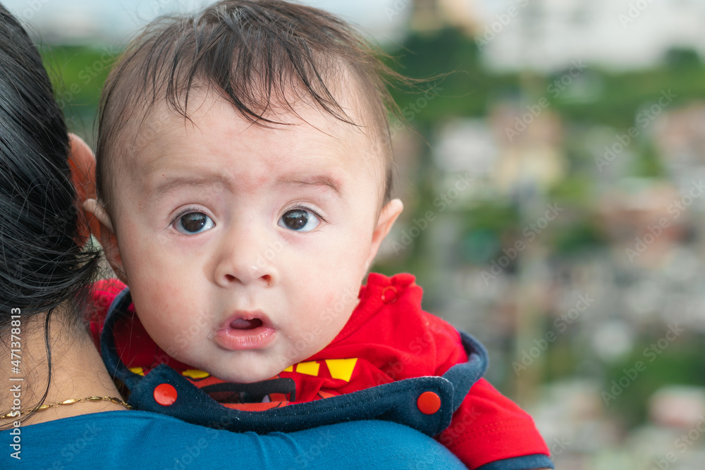 beautiful latin baby peeking his head over his mother's shoulder, big baby with amazed face, blue overalls and red shirt looking back. with his mouth open and big cheeks. concept of motherhood. beautiful latin baby peeking his head over his mother's shoulder, big baby with amazed face, blue overalls and red shirt looking back. with his mouth open and big cheeks. concept of motherhood.