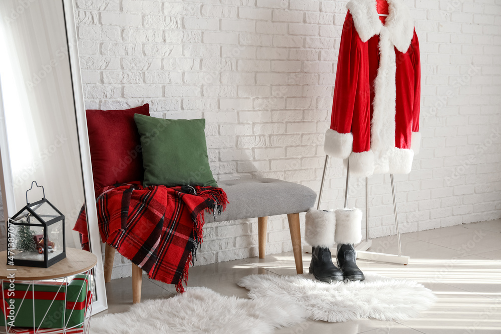 Obraz premium Santa claus costume with boots and christmas decorations in a festive room setting, a santa claus costume with red and white fur trim hangs on a stand, accompanied by black boots with white fur cuffs