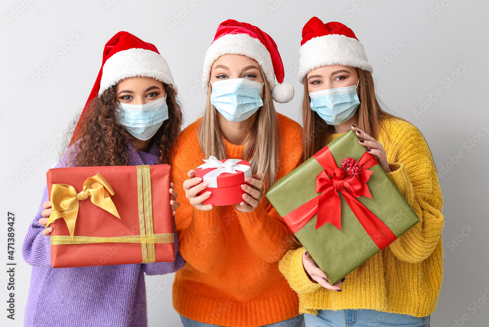 Beautiful young women in medical masks and with Christmas gifts on light background Beautiful young women in medical masks and with Christmas gifts on light background