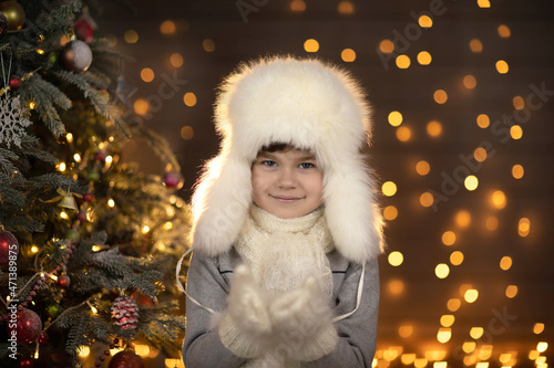 A cute boy in a fur hat smiles and looks at the camera against the background of golden lights, near the Christmas tree.